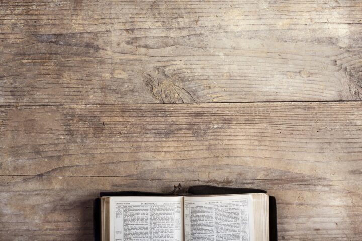 Open Bible on a weathered wooden table with two-page spread of text visible in a black cover case nearby.