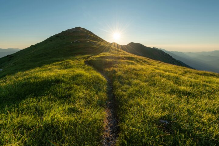 Sunrise over a grassy mountain ridge with a dirt trail leading toward the peak.