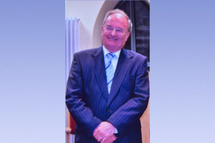 Smiling man in a dark pinstriped suit and blue striped tie, standing with hands clasped indoors.