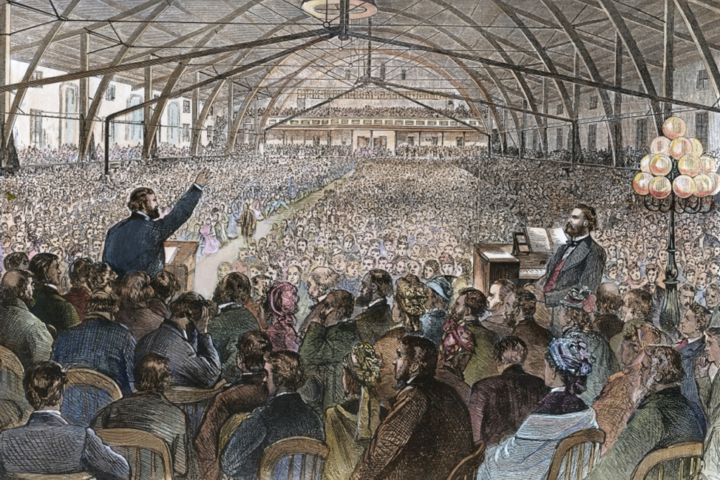 A 19th‑century lecture hall filled with a standing speaker at a podium addressing a huge audience, with a pianist on the right and a vaulted wooden ceiling above.