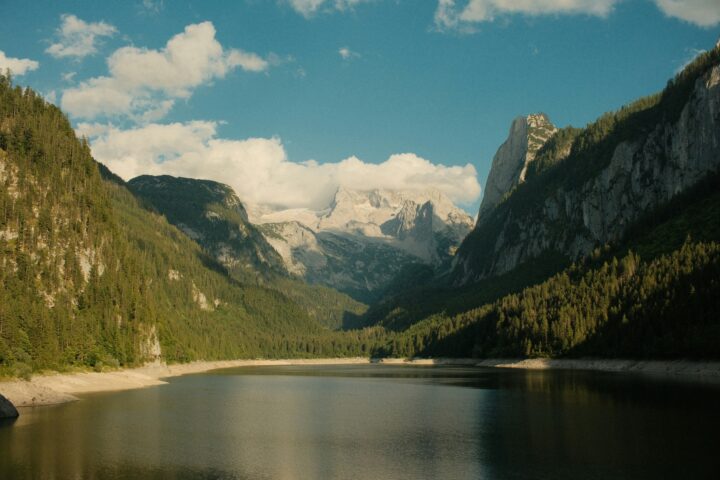A tranquil mountain lake flanked by forested slopes, with snow-capped peaks under a blue sky and scattered clouds.