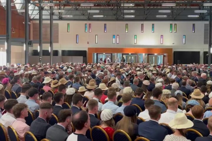 Large indoor conference hall filled with hundreds of attendees seated in rows, many wearing hats, facing a stage at the front.