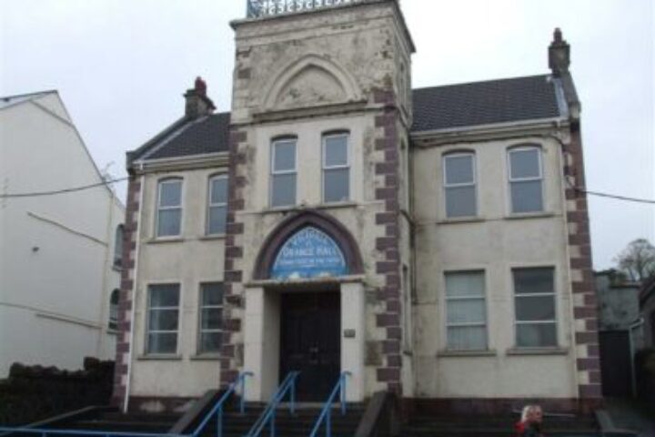 Two-story stone building with a central tower and arched entrance, blue railings along the steps.
