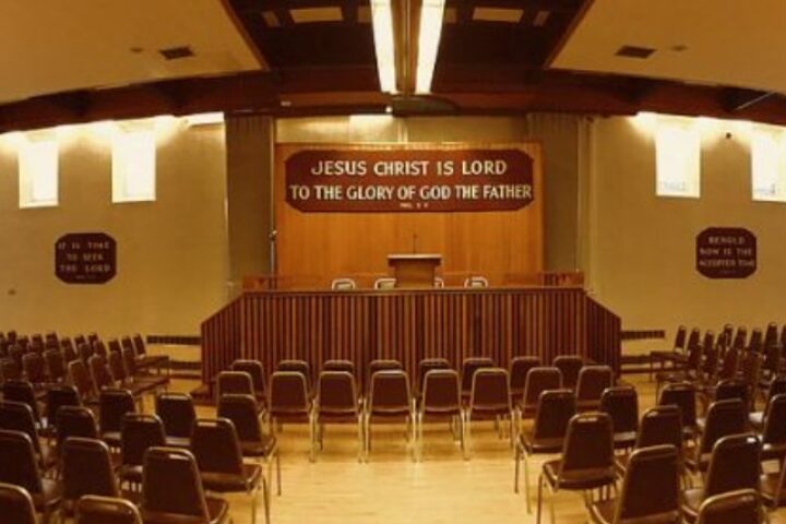 Wide view of a large church sanctuary with rows of brown chairs facing a wooden pulpit and a banner reading 'Jesus Christ is Lord to the Glory of God the Father'.
