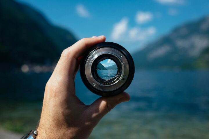 Close-up of a hand holding a camera lens, reflecting mountains and a lake in the lens surface.