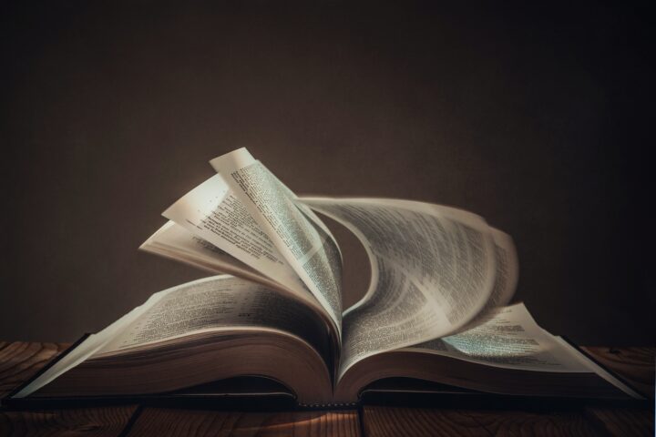 Open books with pages fluttering upward, forming looping curves against a dark background and wooden surface beneath.
