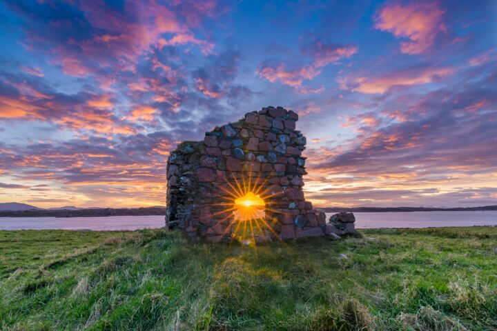 Stone ruin arch with sun bursting through a hole, set on grassy shore above a lake at sunset with colorful sky