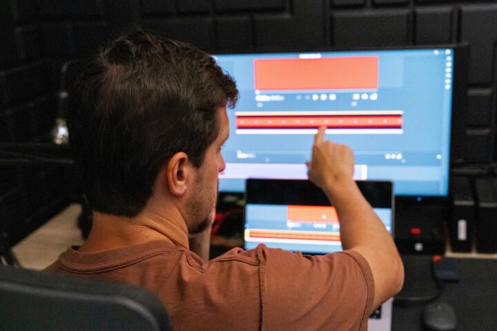 Person shown from the back points at a widescreen monitor displaying a video editing timeline with red and blue blocks.